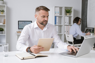 Confident businessman with tablet sitting by desk in front of laptop