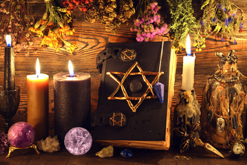 Evil book with pentagram, buning candles, crystal ball, elixir and herbs on witch table.