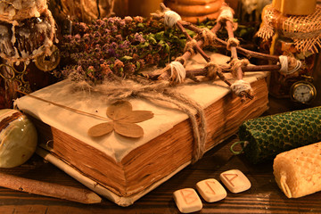 Old book with four-leaf clover, candles, herbs and runes on witch table.