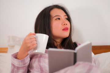 young beautiful and sweet Asian Korean woman in bed reading book novel feeling happy and relaxed or studying at home in cute pajamas cozy and homey drinking coffee