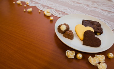 Heart shape cookies, chocolate and chocolate brownie on a white plate with lace and pearl background on brown table.