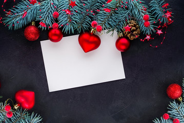 Christmas background: green spruce branches, red toys and a heart-shaped toy, white card on a black textured background. Template for design, greeting card, greeting.Top view. Flat lay