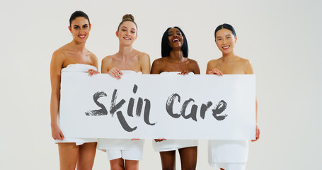The beautiful young smiling women of different ethnicities with perfect firm and slim body in towels keeping a cartel with "Skin Care" and smiling in camera. 