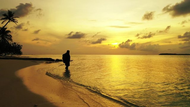 Golden sunset with yellow sun setting down the grey clouds, reflecting over calm glassy surface of sea washing pristine beach with bent palms, silhouette of girl 