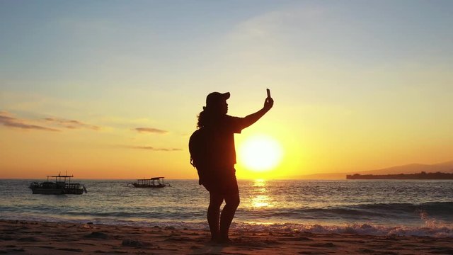 Girl taking photos with her smartphone on beautiful exotic beach washed by sea waves at sunset with dusty yellow sky over tropical bay