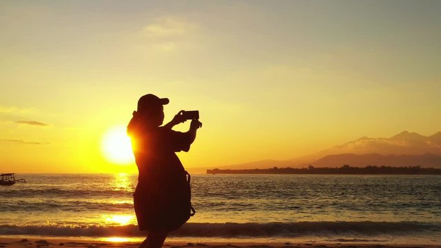 Silhouette of young woman taking photos of beautiful sunset with yellow misty sky reflecting on sea surface near shore of tropical island 