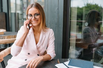 Fototapeta na wymiar Smiling businesswoman dressed in formal wear having pleasant conversation on mobile phone with friend, happy attractive female speaking on cell telephone while resting in office interior after work