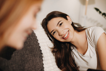 Portrait of a lovely young woman with long dark hair laughing while leaning head on the sofa looking at her female friend with red hair.