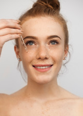 Close up portrait of a young beautiful red haired woman applying hyaluronic acid with a dropper smiling isolated on a white background.