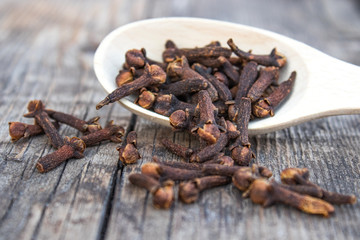 A spice of dried cloves lies on a wooden spoon and is scattered on old wooden boards.