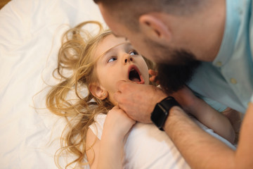Cute little kid leaning on a bed with mouth opened while her father is looking at her teeth. Young father looking in the mouth at his daughter at the teeth.