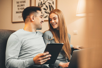 Beautiful young couple smiling face to face sitting on a couch at home and using a tablet and a laptop.