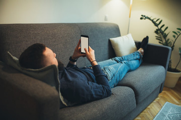 Bearded young man leaning on the couch and looking at smartphone screen at his home after work.