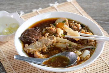 Chinese roll noodle in dark soup with crispy pork in white bowl and chili pickled souce in white cup all put on bamboo mat