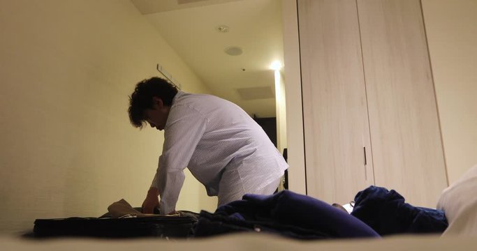 From below side view of female in white pajama putting things to bag while preparing for sleep in small hotel room with minimalist interior with beige walls and wardrobe