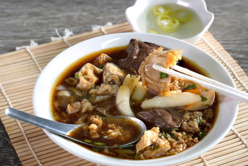 Chinese roll noodle in dark soup with crispy pork in white bowl and chili pickled souce in white cup all put on bamboo mat