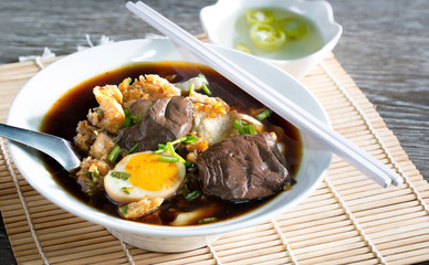 Chinese roll noodle in dark soup with crispy pork in white bowl and chili pickled souce in white cup all put on bamboo mat