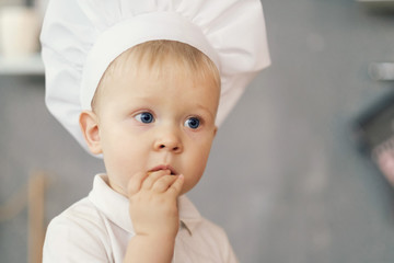 one little boy in hat of chef in kitchen sitting on the table and eat