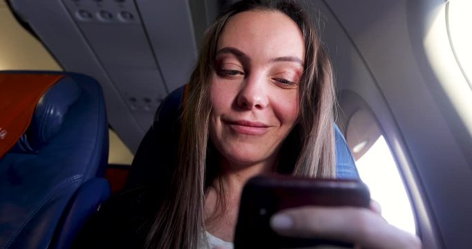Happy young female passenger sitting in airplane next to window and checking messages on smartphone before flight