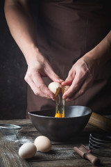 Professional chef hands are breaking an egg into bowl to make dough on wooden table, over dark background