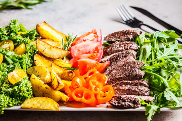 Chopped beef steak, baked potato wedges and vegetables on gray plate, gray background.