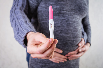 Young pregnant woman and her husband holding swollen belly with pregnancy test in his hand.
