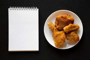 Homemade crispy oven baked fried chicken on a white plate, blank notepad over black background, top view. Copy space.