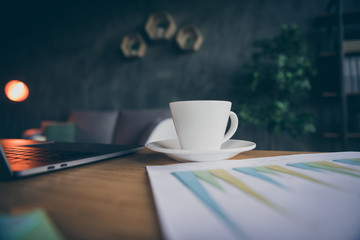 Cropped close up photo of cup of coffee ready to be drunk standing on plate near diagram work paper and laptop to the right