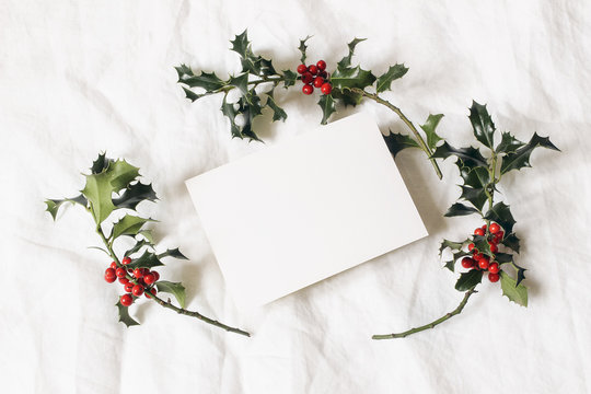 Christmas styled stock photo. festive greeting card, invitation mockup scene. Still life with paper card and holly berries on white linen background. Winter stationery composition. Flat lay, top view.