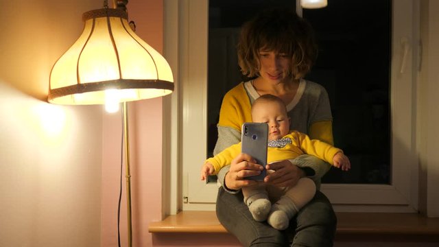Mother with baby child enjoy take phone selfie pictures sitting on windowsill near lamp at home