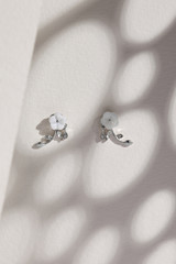 Subject shot of a pair of stud earrings isolated on the beige surface with gray shadows. Each of the earrings is made as a white flower, silver leaves and crystals. 