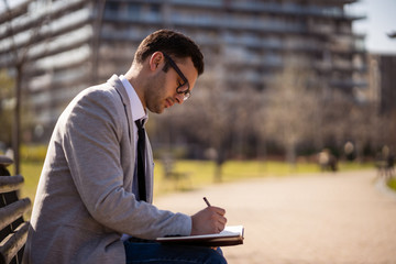 Young businessman is sitting in park and making plans.