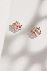 Subject shot of a pair of stud earrings isolated on the beige surface with gray shadows. Each of the earrings is made as a golden flower crown with shimmering white pearls, gems and crystals. 