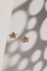 Subject shot of a pair of stud earrings isolated on the beige surface with gray shadows. Each of the earrings is made as a golden star with sparkling crystals, gem flowers and jeweller's enamel.