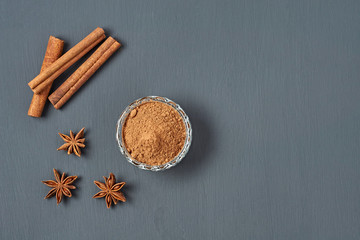 Three cinnamon sticks near glass bowl full of powder and scattered stars of anise lies on dark scratched desk on kitchen. Space for text. Top view