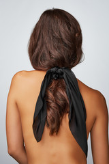 Fototapeta premium Close-up back shot of a brunette lady with a black silk hair scarf fixed on the scrunchie. The shirtless woman with a ponytail is posing on the gray background. 