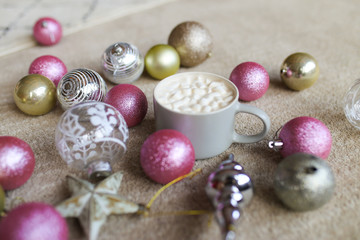 Fototapeta na wymiar Christmas picture. A white Cup of cocoa or coffee with marshmallows stands among silver, gold and pink Christmas balls, toys, stars and decorations on a beige background