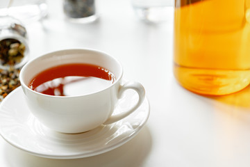 Cup of hot black tea on white table with dry tea jar