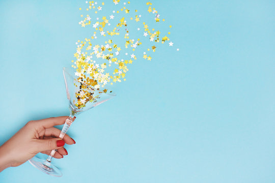 Woman hand holding martini glass with pouring out golden stars confetti on blue background.