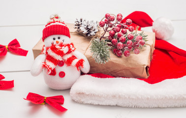 Toy snowman with Christmas present and red hat on a white wooden table.