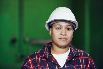 Head and shoulders portrait of mixed-race woman wearing hardhat looking at camera while posing in industrial workshop against green wall, copy space