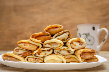 Pancakes with filling are a pyramid on a white plate standing on a wooden background close-up, in the background a Cup