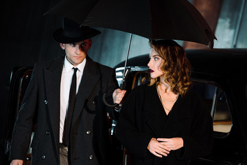 Naklejka premium handsome man in hat holding umbrella near attractive girl and vintage car