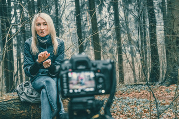 blonde girl in a blue jacket, jeans sits in the forest on a fallen tree and writes a blog on camera, showing emotions of surprise, waving her hands. concept of blogging, modern technology.