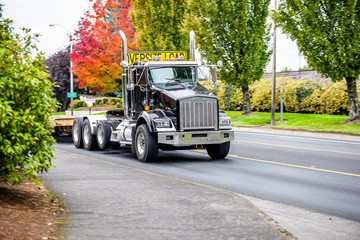 Fototapeta na wymiar Powerful big rig black semi truck with step down semi trailer and sign for transporting oversize load running on the local road with autumn trees on the side