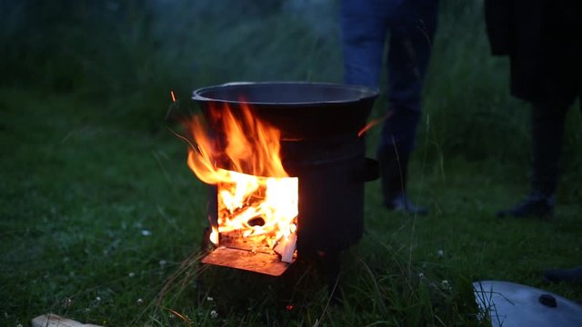 Cauldron on the river bank. A fire burns under it, firewood sticks out. Evening, fishermen are going to cook fish soup