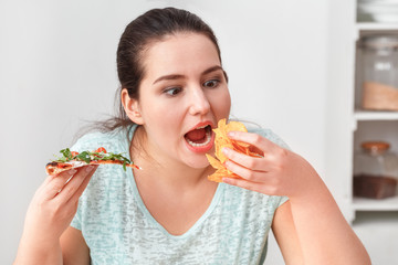 Binge Eating. Chubby girl sitting at kitchen table eating pizza and chips hungry close-up