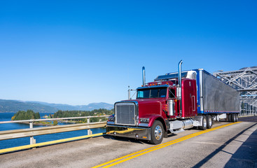 Red classic bonnet big rig semi truck transporting frozen cargo in bright refrigerated semi trailer...