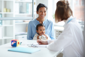Smiling young mother sitting together with her baby and talking to the doctor during their visit at...