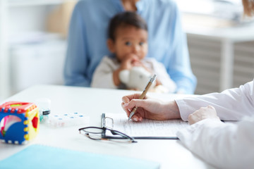 Close-up of female pediatrician sitting at the table and writing a prescription with mother with...